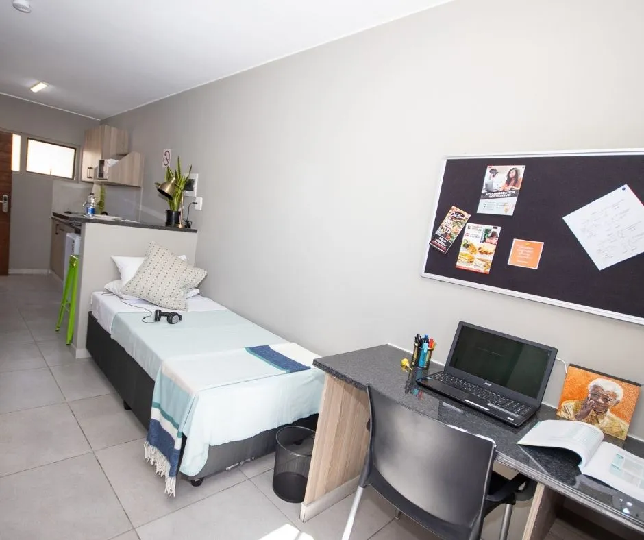 A furnished single room with a bed, desk and pin board